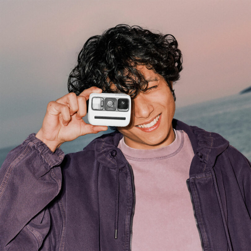 Person smiling and holding a compact camera up to their eye, outdoors with a pastel sky in the background.