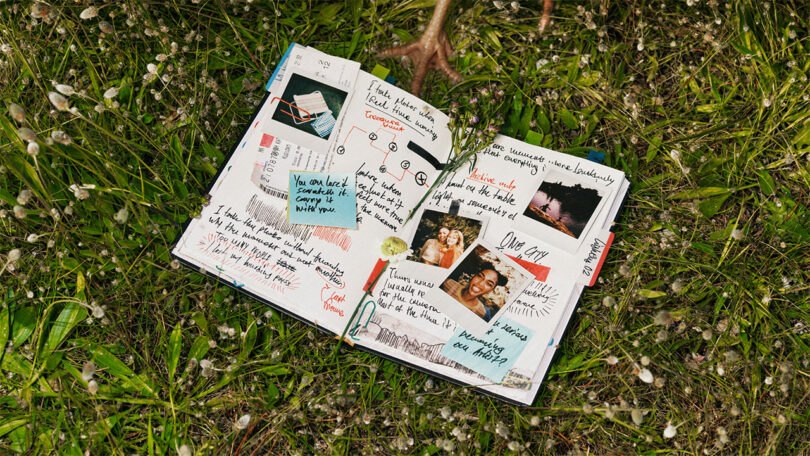 An open scrapbook with handwritten notes, photos, and doodles lies on green grass with small white flowers.