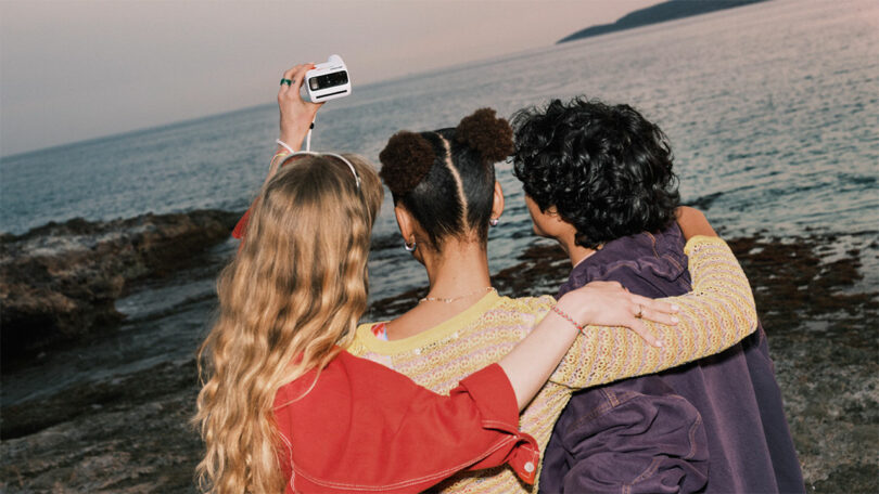 Three people with their backs to the camera stand by the rocky shore, one holding up an instant camera to take a photo with the sea and sky in the background.