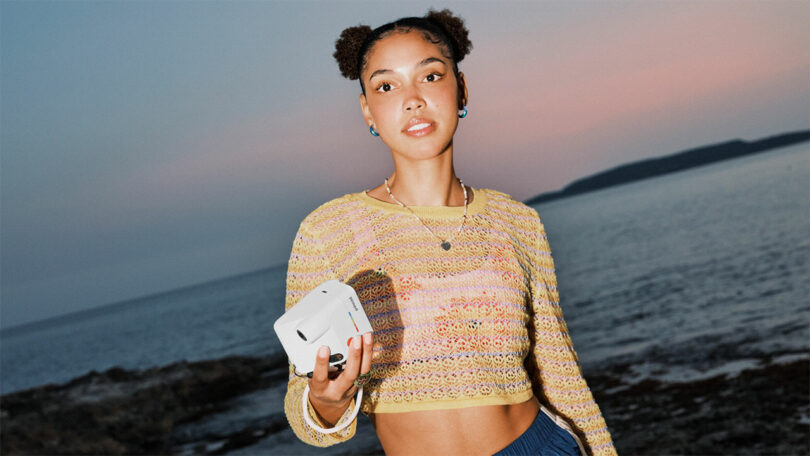 Young woman with two buns holding a white camera stands near the seaside at dusk, wearing a yellow crochet crop top and blue pants.