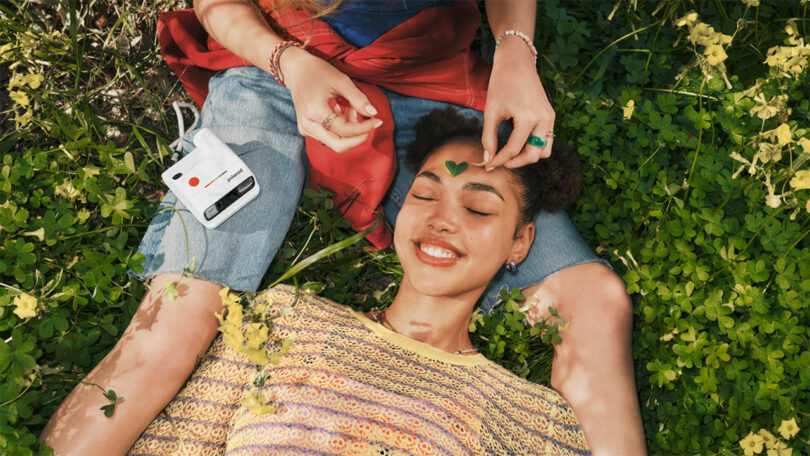 A person lies smiling on grass with their head in another's lap. One hand places a green leaf on their forehead; a white instant camera rests nearby.