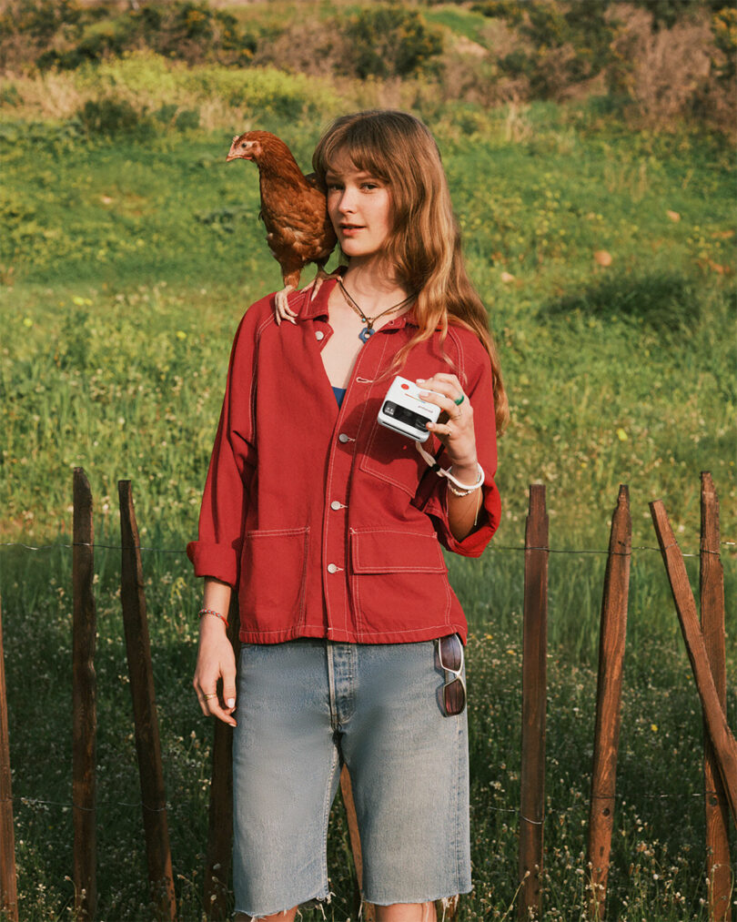 A person in a red shirt and denim shorts stands outdoors holding a camera, with a brown chicken perched on their shoulder. Green grass and wooden fence are in the background.