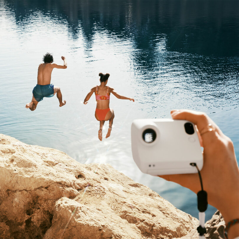 Two people jump off a rocky ledge into a lake while a hand in the foreground holds a camera, capturing the moment.