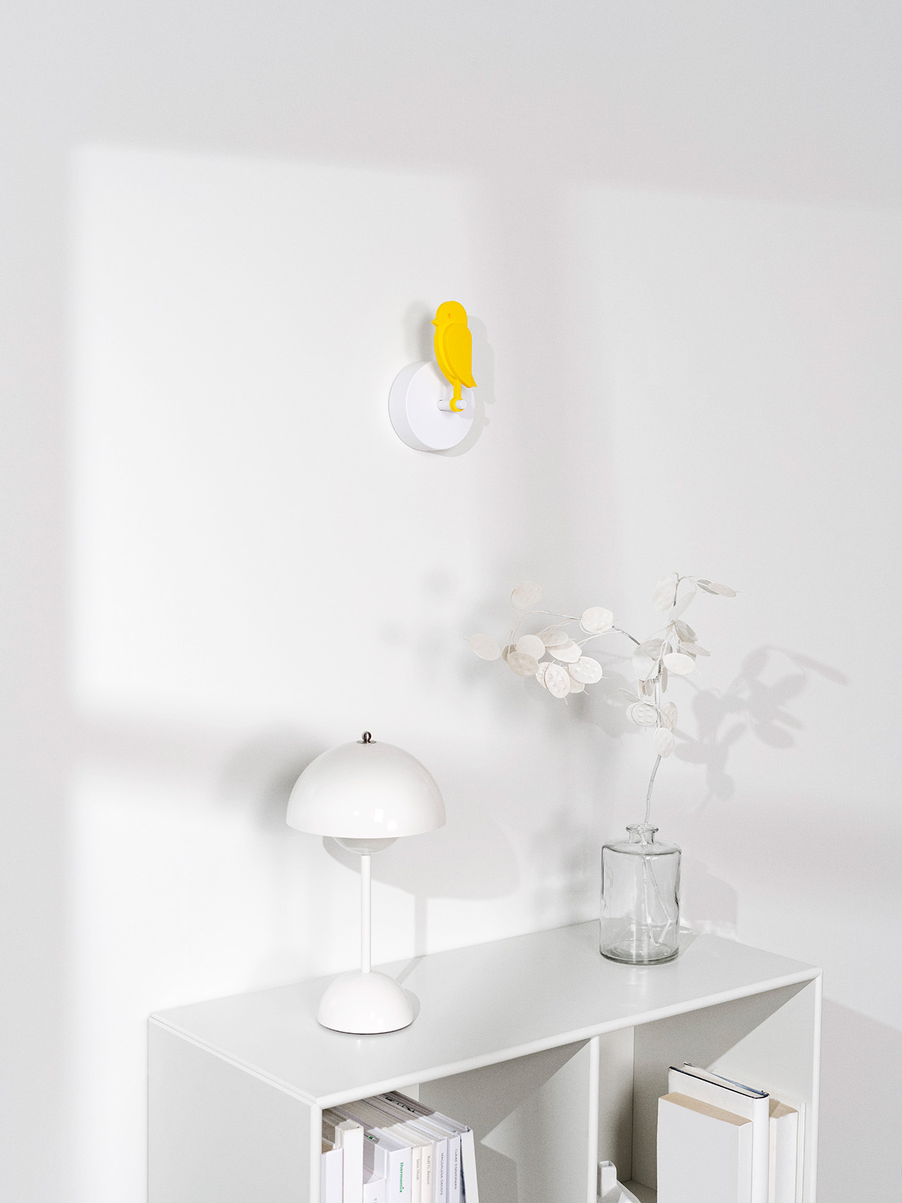 Una estantería blanca, una lámpara de mesa blanca, un jarrón de cristal con hojas blancas y una alegre decoración de pared en amarillo Birdie complementan las paredes blancas con tonos suaves.