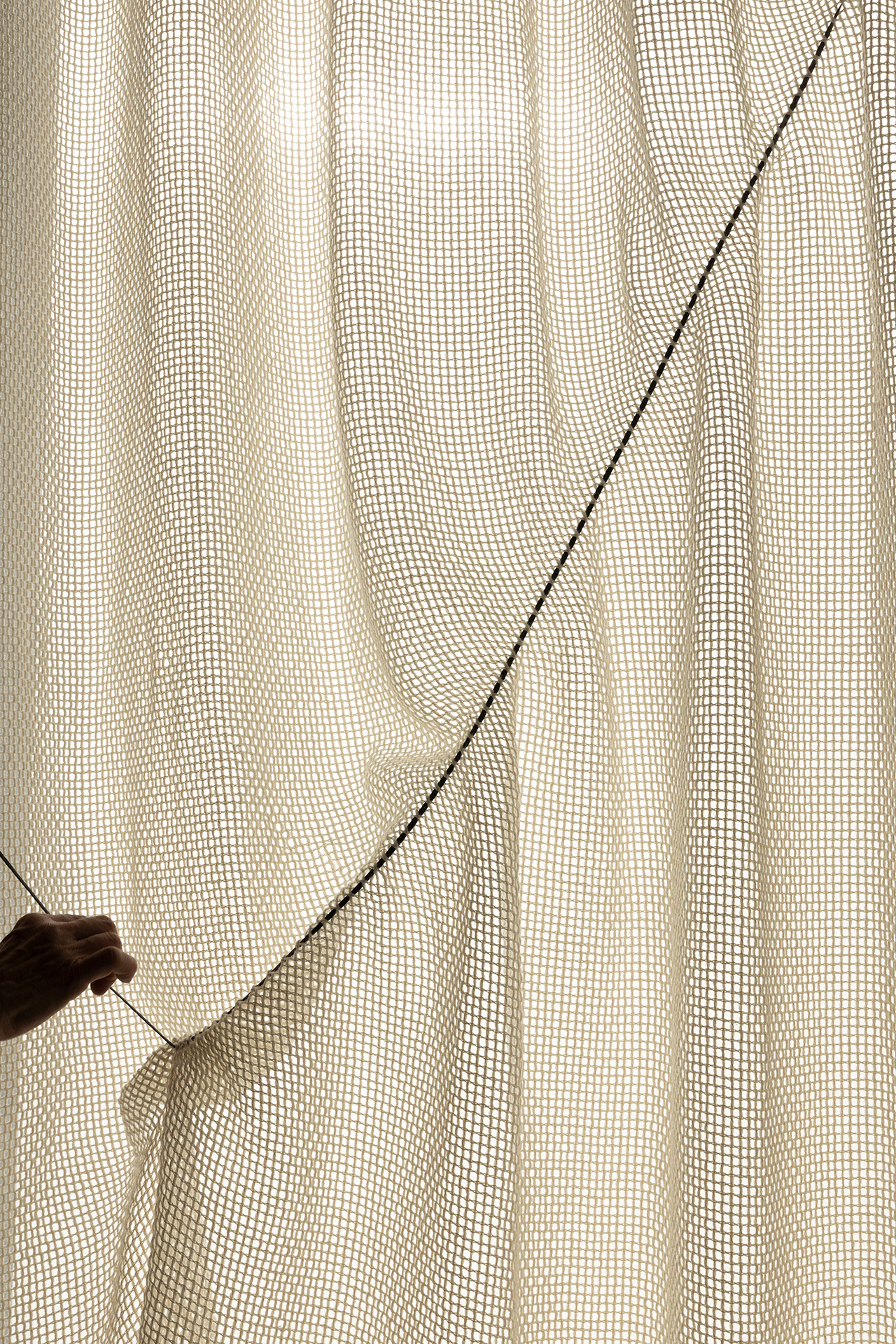 A hand pulls back a sheer, textured curtain revealing light shining through its woven pattern.