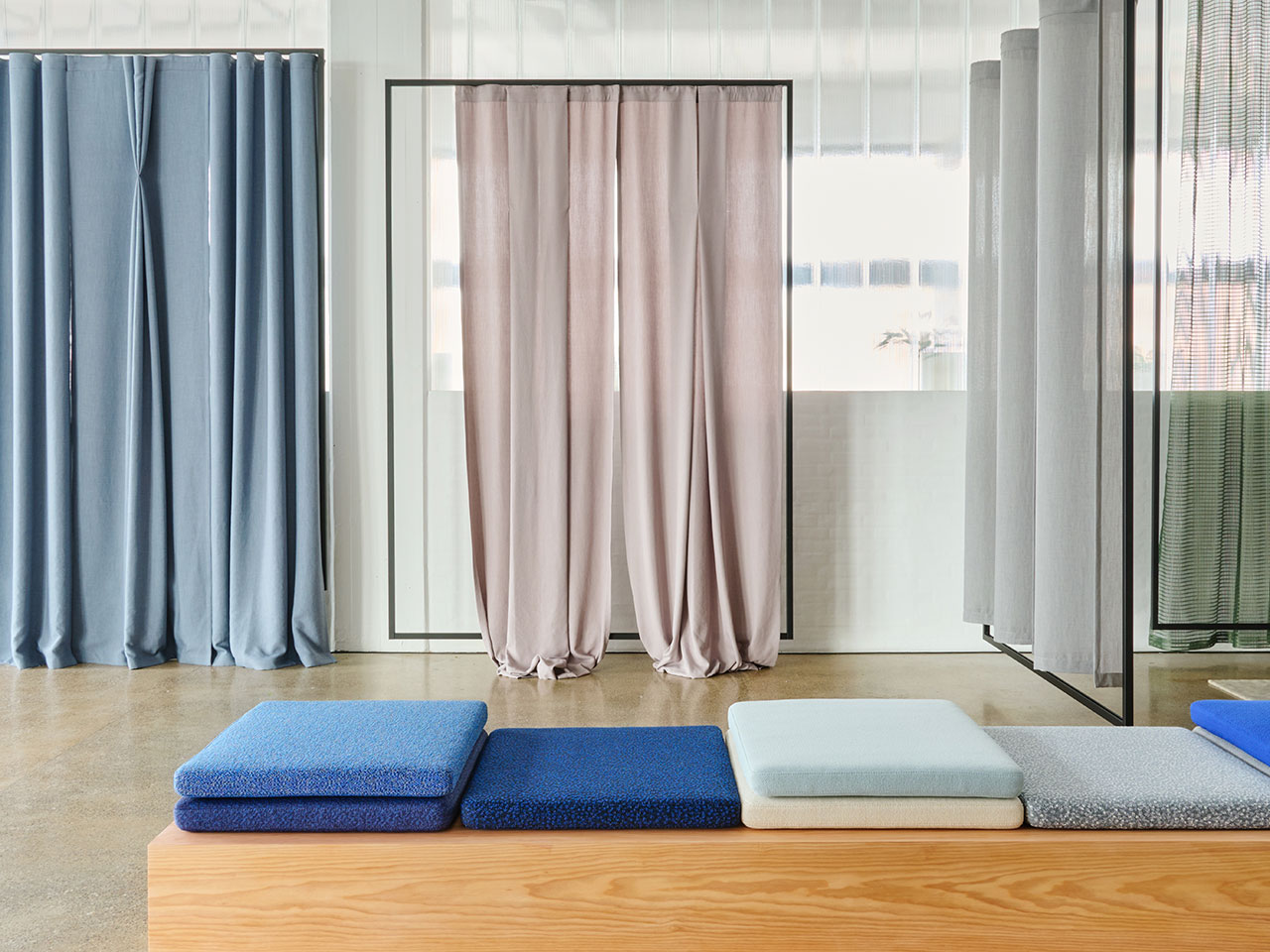 A wooden bench with blue and gray Kvadrat seat cushions sits in front of floor-length curtains in blue, beige, and gray tones. The floor is polished concrete.