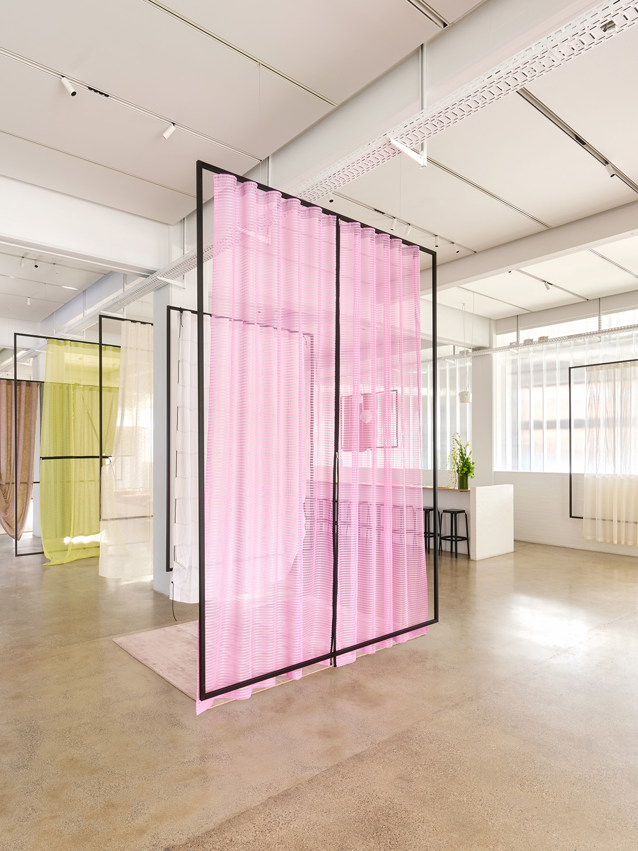 Modern interior space with translucent colored fabric panels, including pink, yellow, and beige, hanging from black metal frames on a polished concrete floor.
