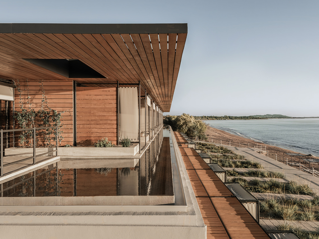 Modern wooden building at Dexamenes with a reflecting pool overlooks a sandy beach and calm sea under a clear sky.