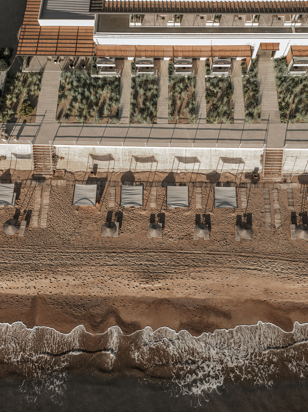 Aerial view of a sandy beach at Dexamenes, with rows of sun loungers and umbrellas near the water, bordered by a walkway and lush vegetation.