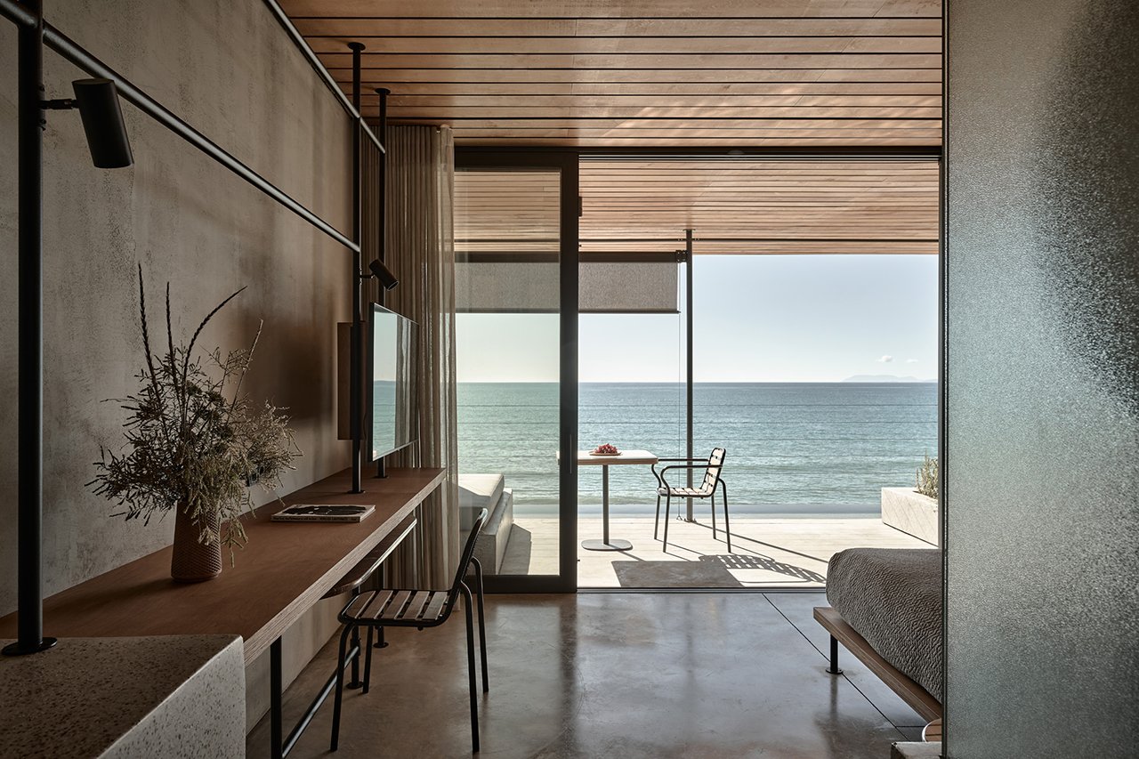 Minimalist Dexamenes beachfront hotel room with a desk, chair, bed, and sliding glass doors opening to a patio with a small table and two chairs overlooking the ocean.