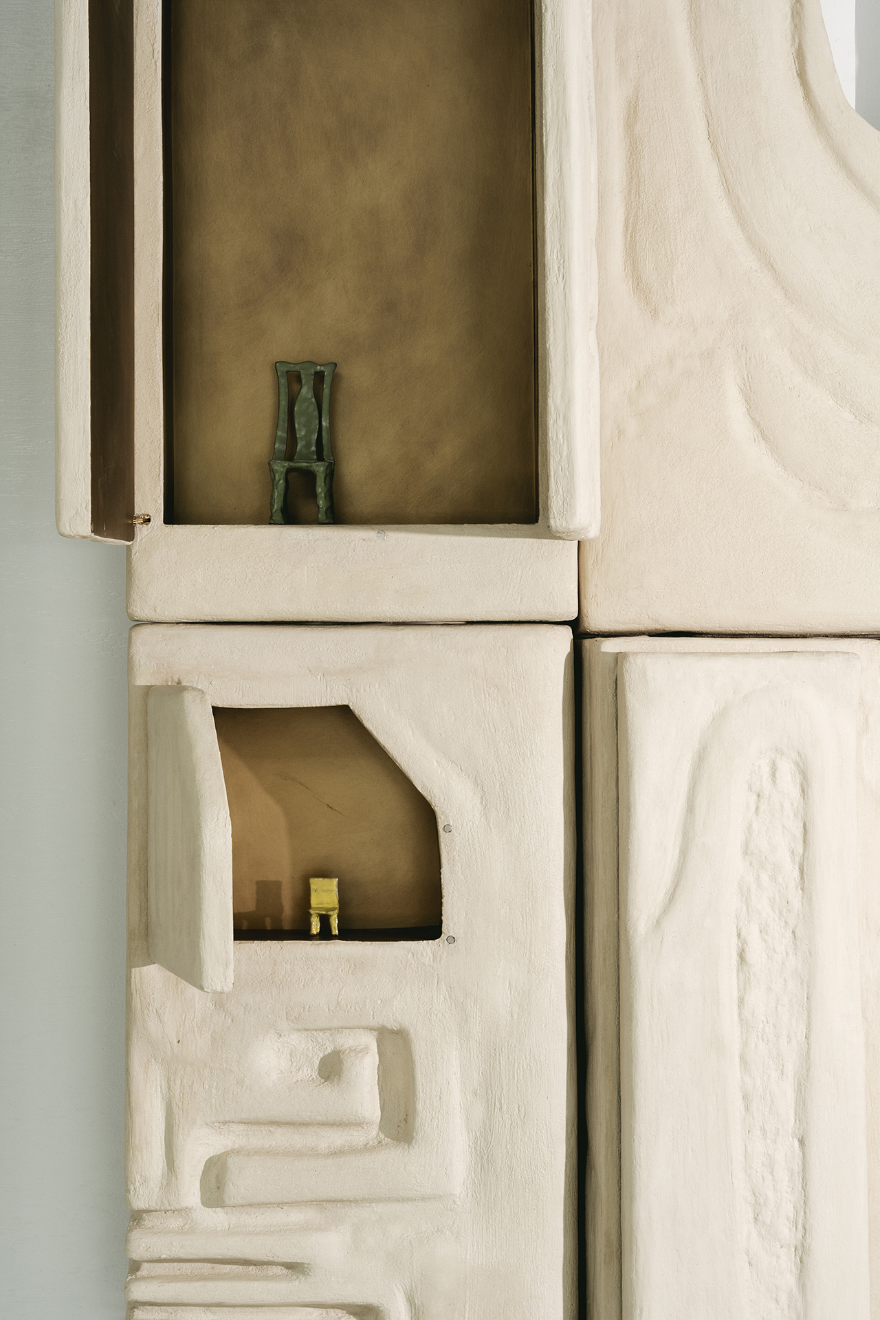 Two small shelves built into a textured, off-white wall hold miniature sculptures—a green chair on the upper shelf and a yellow chair on the lower shelf.