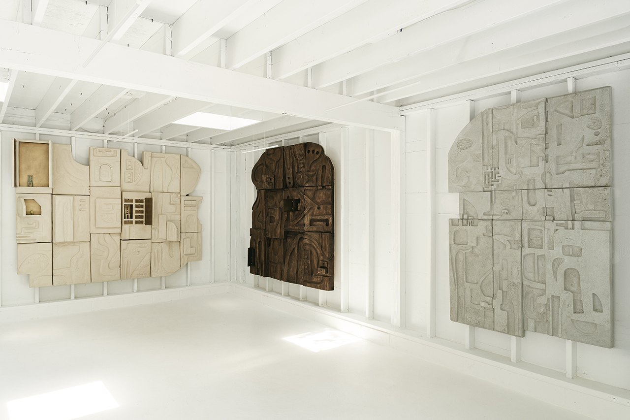 A white gallery room displays three large abstract relief wall sculptures in beige, brown, and gray tones, each composed of geometric and organic shapes.