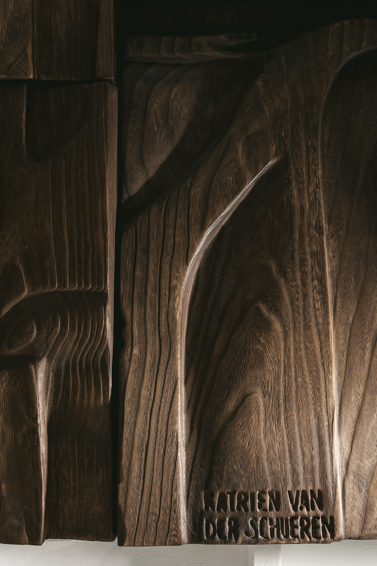 Close-up of carved dark wood panels with abstract patterns and the name "MARIEN VAN DER SCHUEREN" engraved on the surface.