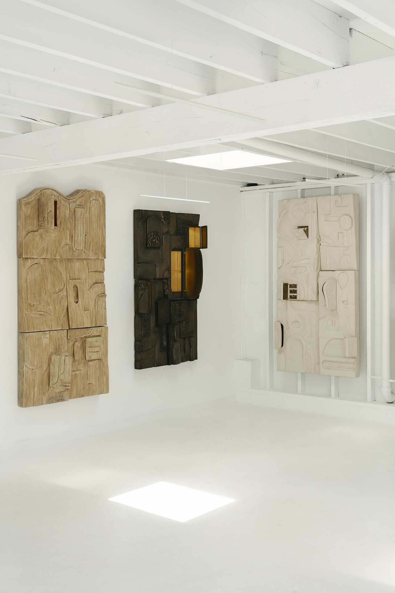 Three large abstract wall sculptures in neutral tones are displayed on a white gallery wall with exposed beams and a white floor.