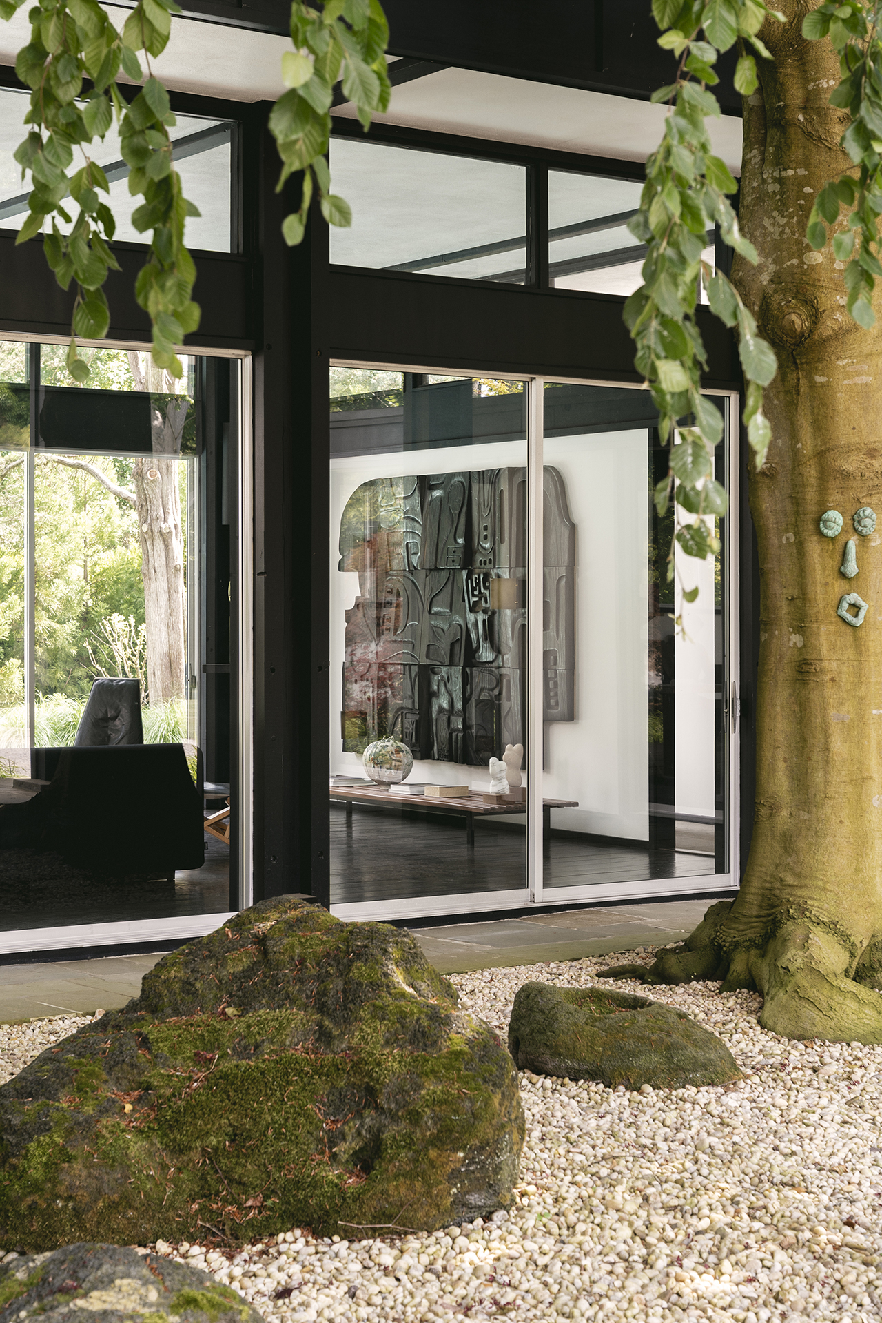 Large glass doors reveal a modern interior with abstract wall art, viewed from a gravel garden with mossy rocks and a tree in the foreground.