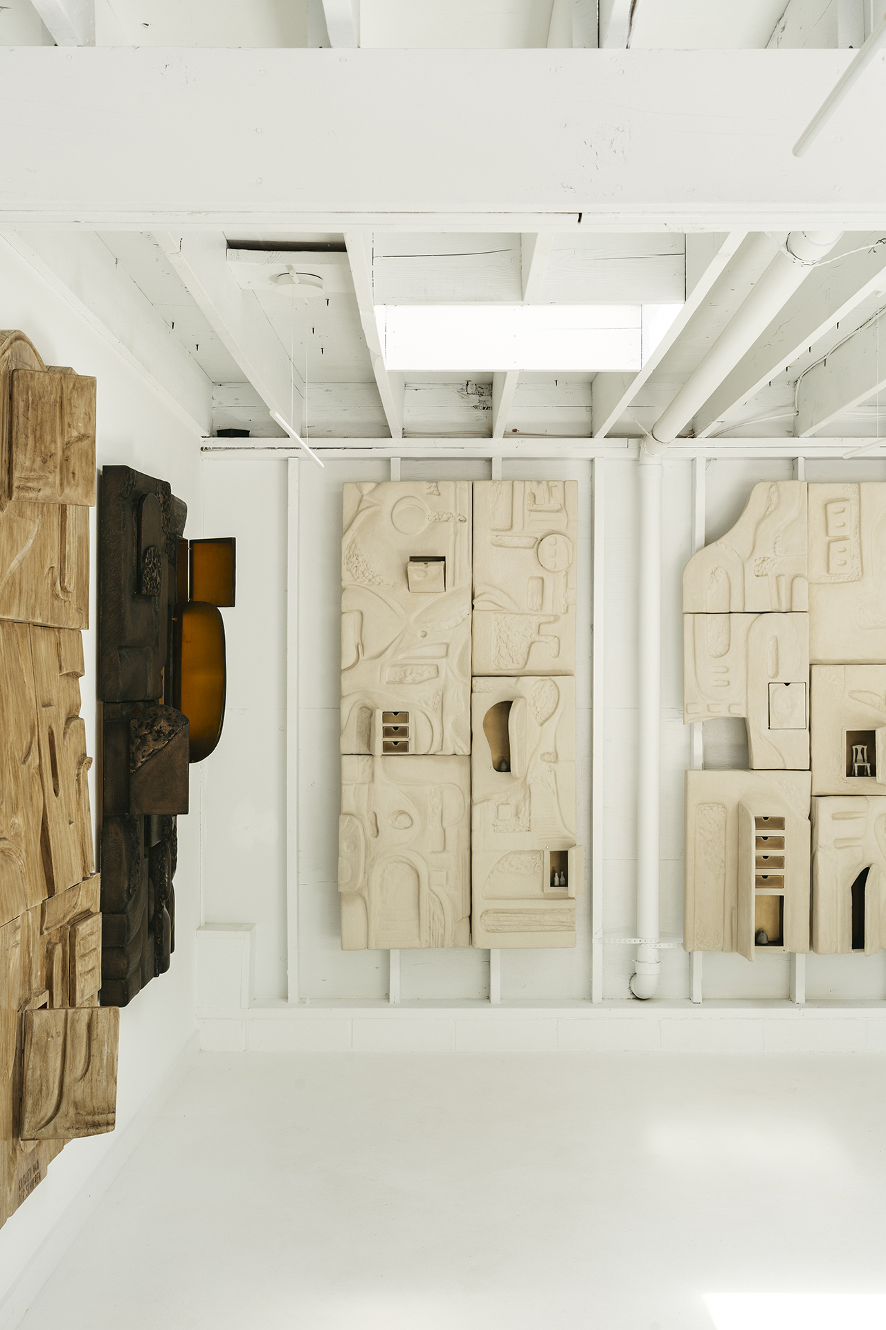 Two large abstract wall sculptures made of light and dark carved materials are displayed in a white gallery space with exposed beams.