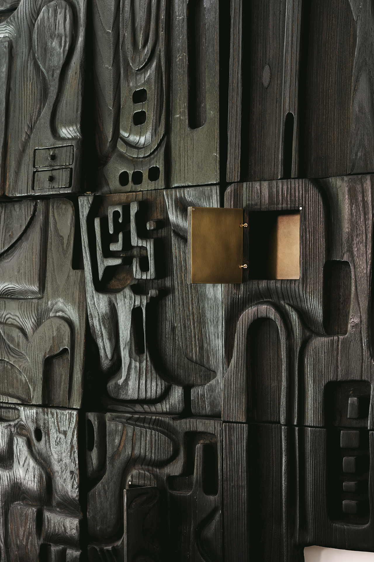 Dark carved wooden wall with abstract geometric patterns and a rectangular brass wall sconce attached near the center.