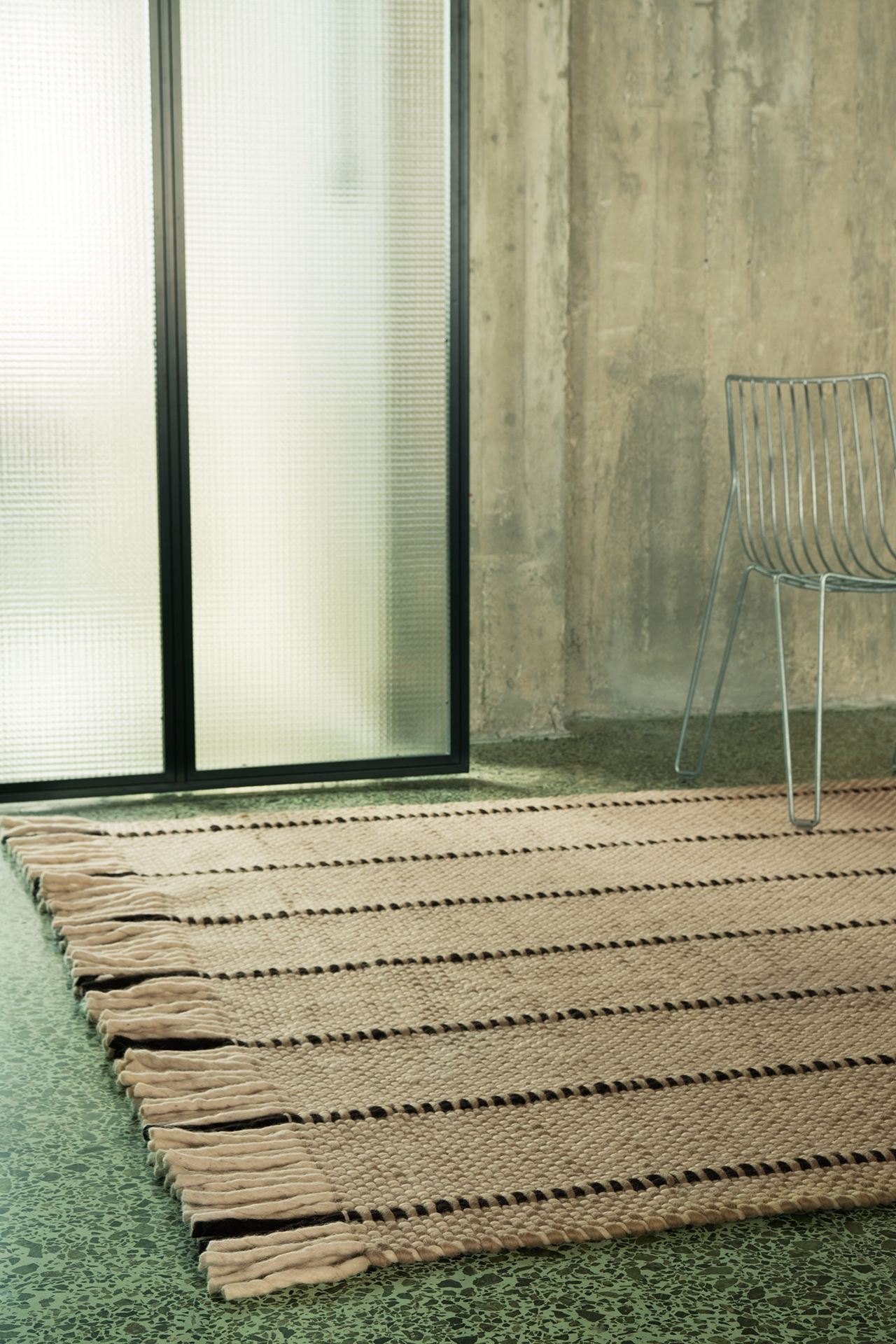 A woven rug with black stripes and fringed edges is layered on a green speckled floor near a metal chair and a frosted glass door in a concrete room.