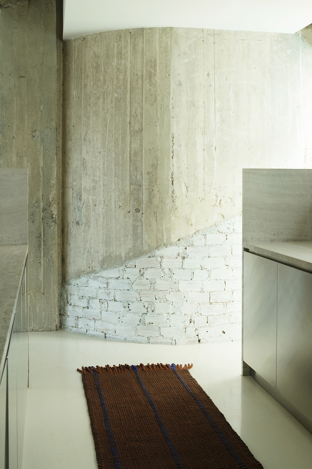 A modern kitchen features a layered mix of rough concrete and white brick walls, white floor, stainless steel counters, and a brown woven rug with blue tassels.