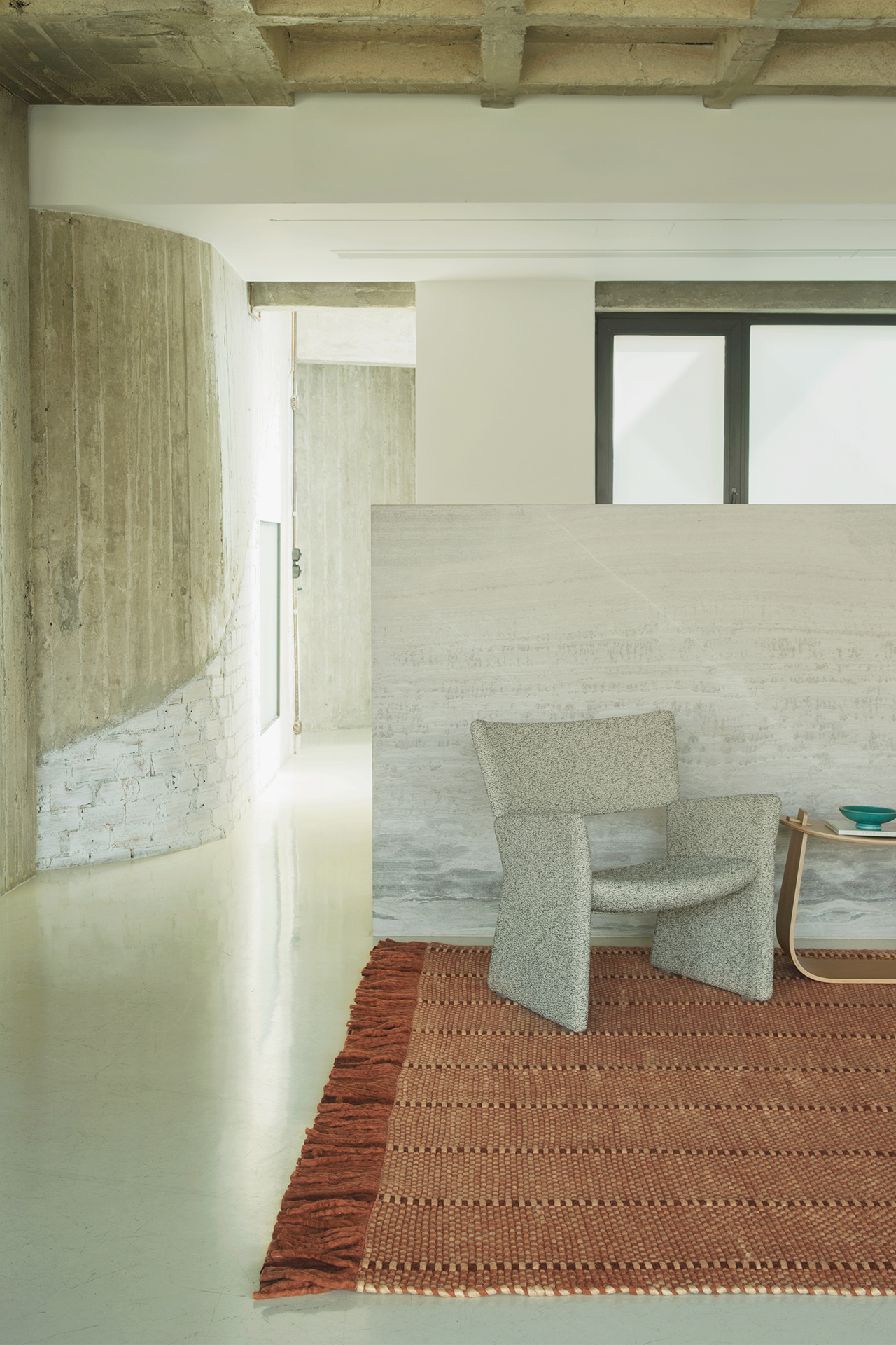 A modern minimalist interior with exposed concrete walls, a gray armchair, and a wooden coffee table creates a LAYERED look, accented by a rust-colored textured rug on a light floor.