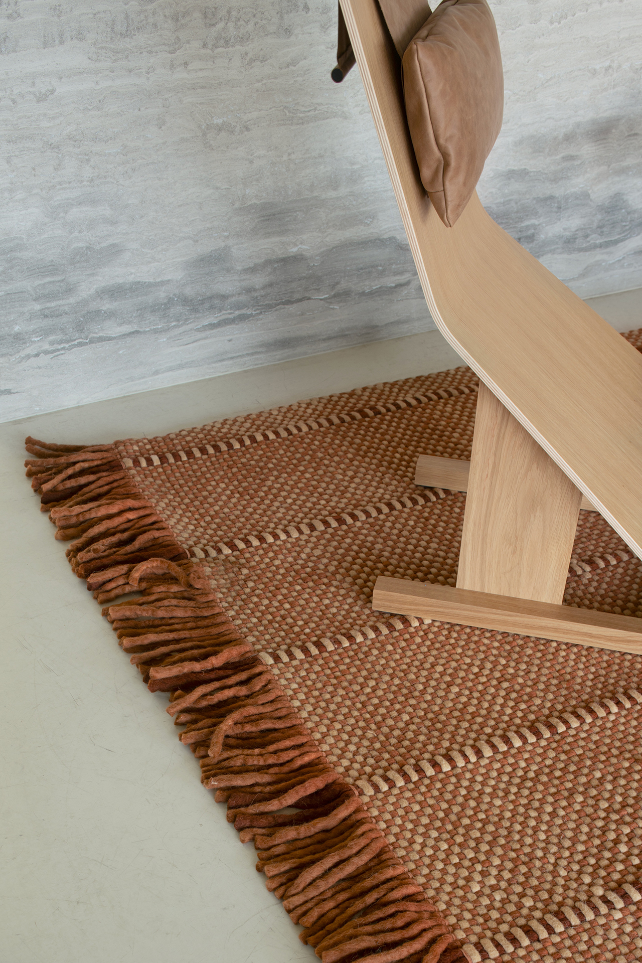 A wooden lounge chair with a brown cushion sits on a woven rug with fringed edges, creating a layered look next to a light-colored wall.