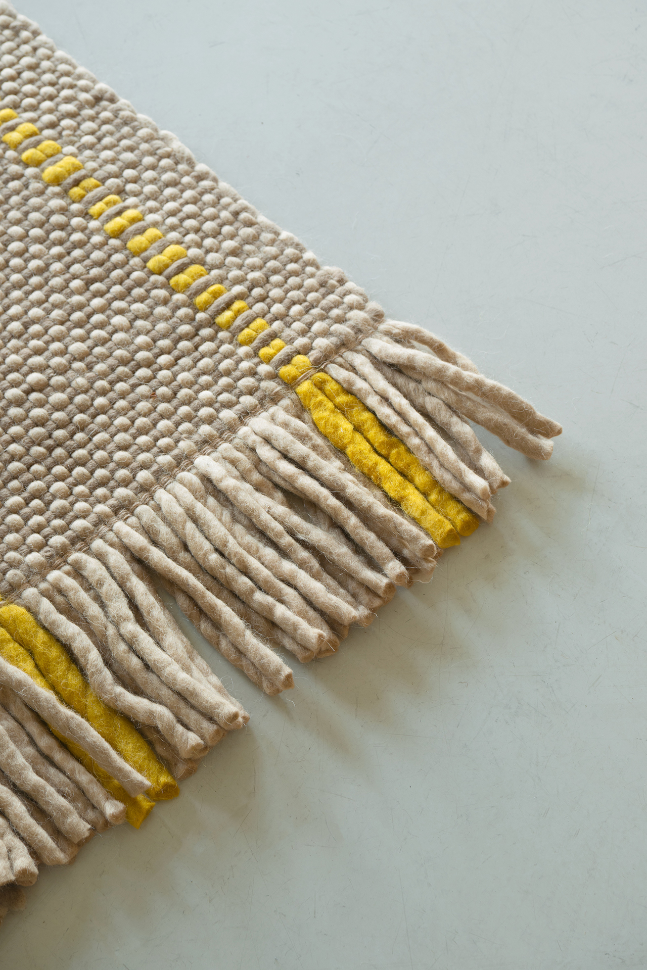 Close-up of a beige woven rug with thick yarn, yellow stripe accents, and fringed edges layered over a light gray surface.