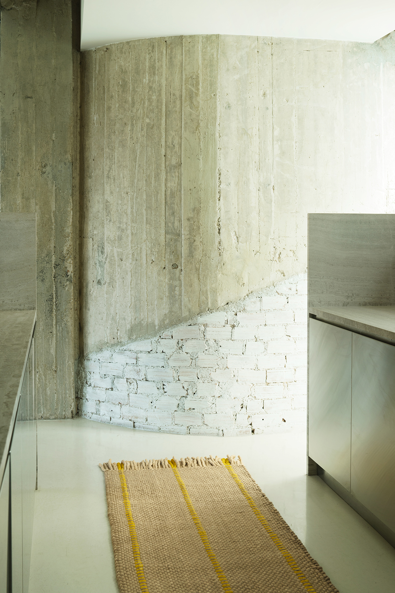 Modern minimalist kitchen with concrete and white brick wall, light-colored floor, stainless steel cabinets, and a layered woven rug with yellow stripes.