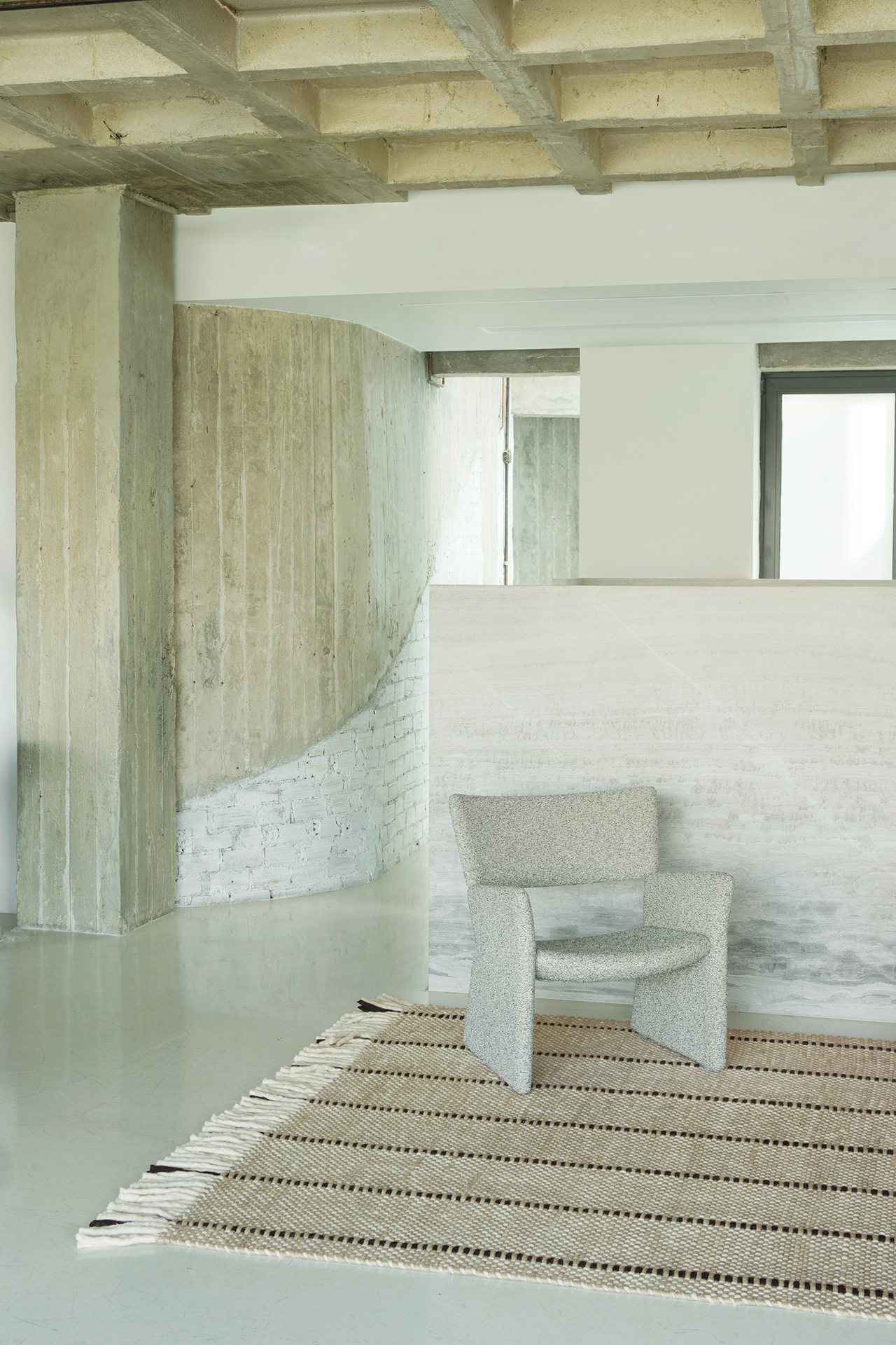 A modern minimalist room with exposed concrete, a layered textured gray chair, a neutral woven rug, and a mix of white and gray walls.