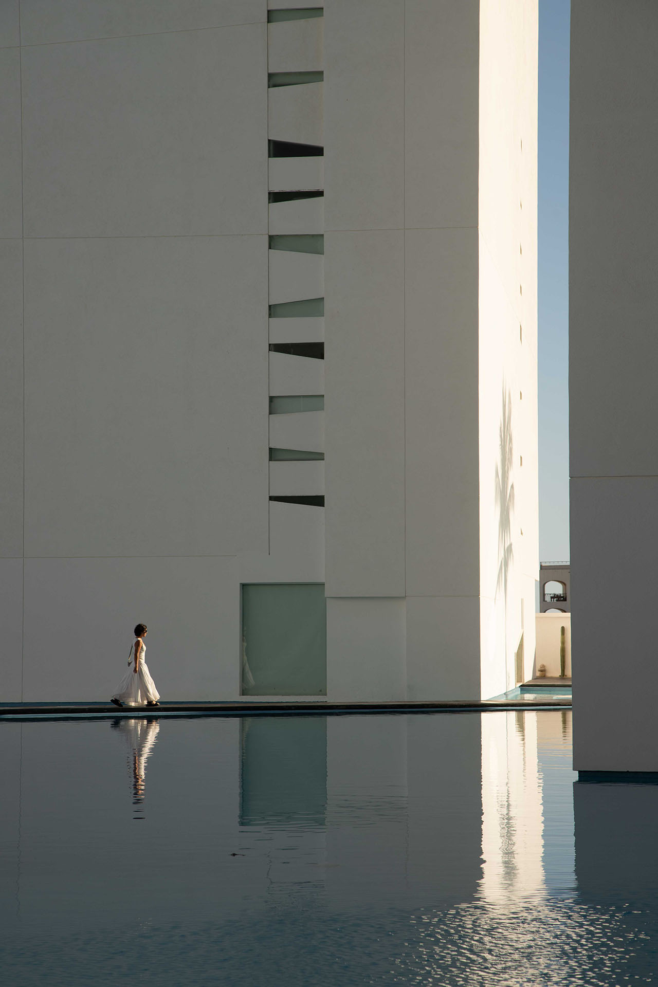 A person in a white dress walks alongside a modern, white building reflected in a still pool of water under clear daylight.