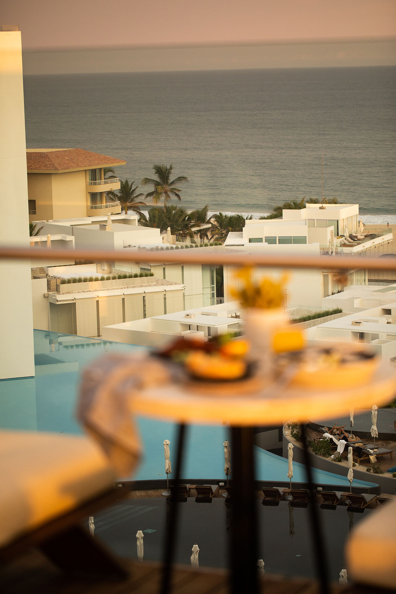 A table with food and drinks is in the foreground, overlooking a resort with a pool, palm trees, and the ocean in the background.