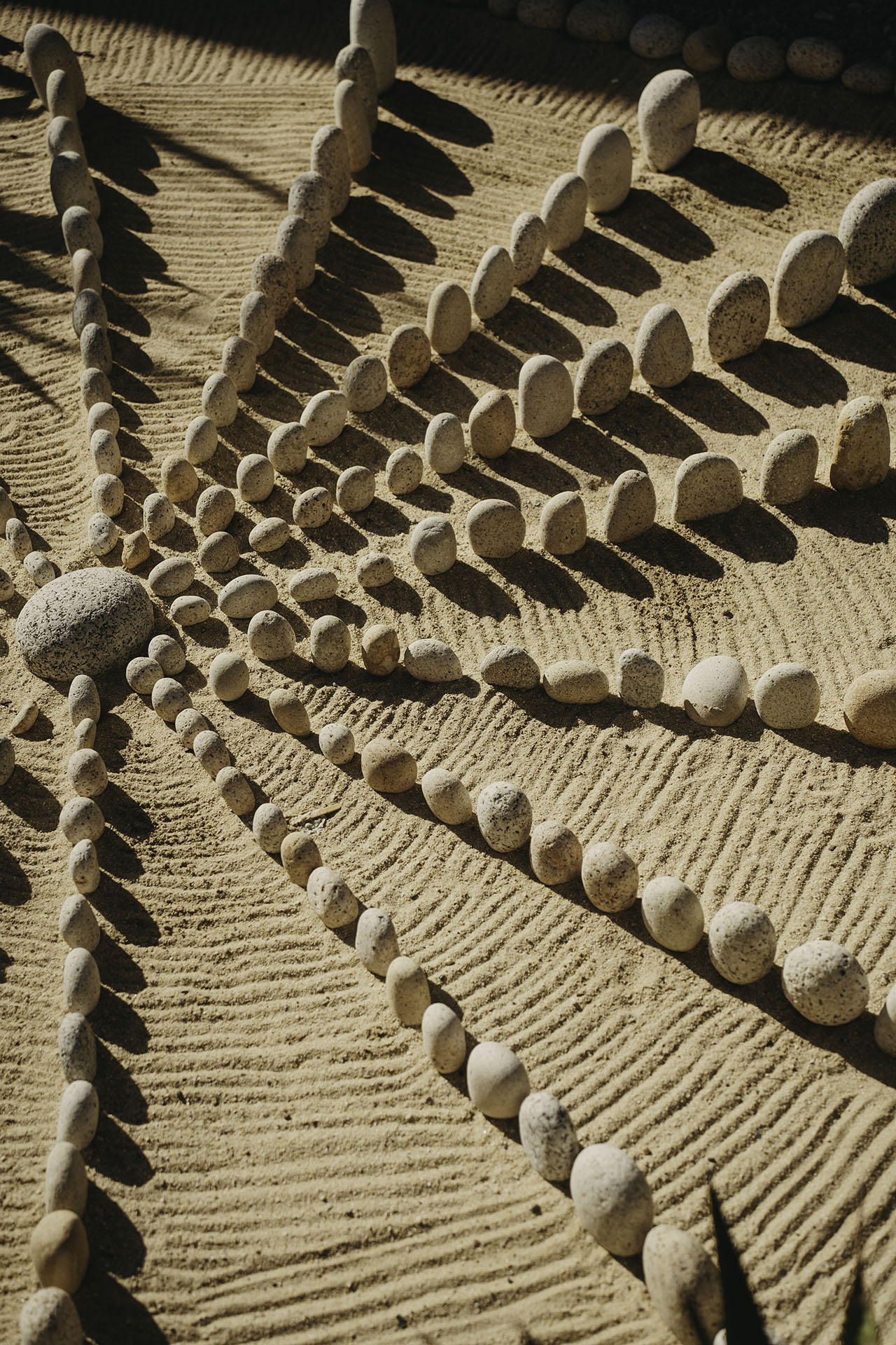 Lines of small stones radiate from a central point on raked sand, creating a symmetrical pattern with shadows and textured lines.