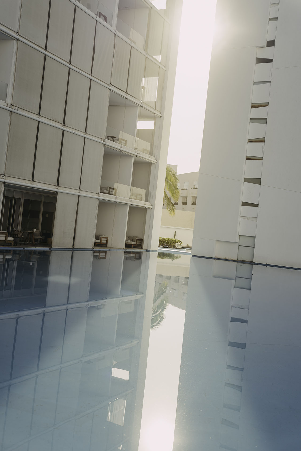 Sunlight shines between two modern white buildings, reflecting off a still, blue swimming pool surrounded by minimalist balconies and loungers.
