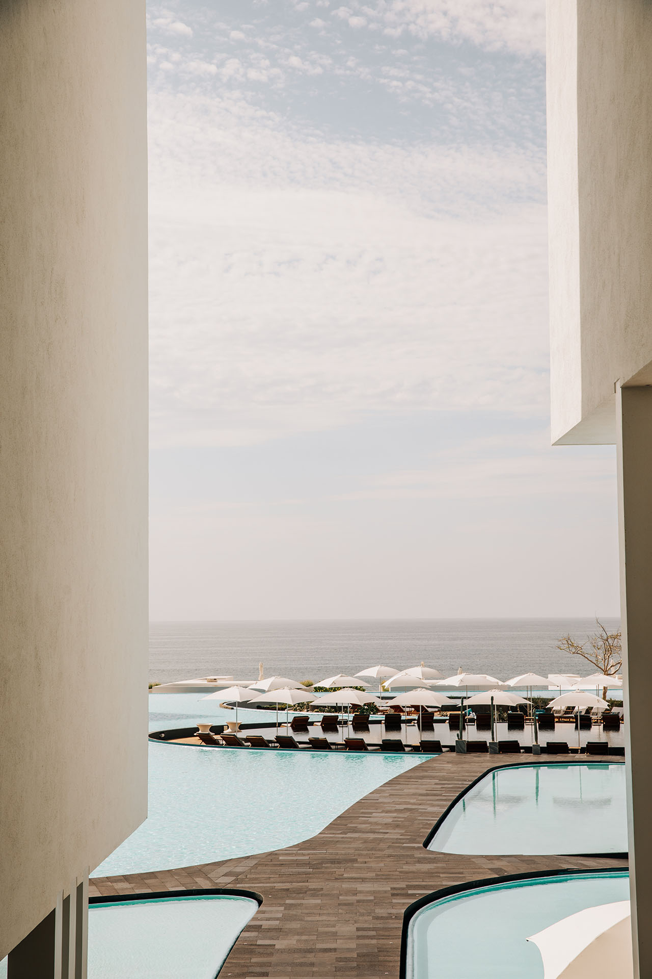 View of a resort pool area with lounge chairs and white umbrellas facing the ocean, framed by two white buildings, under a partly cloudy sky.