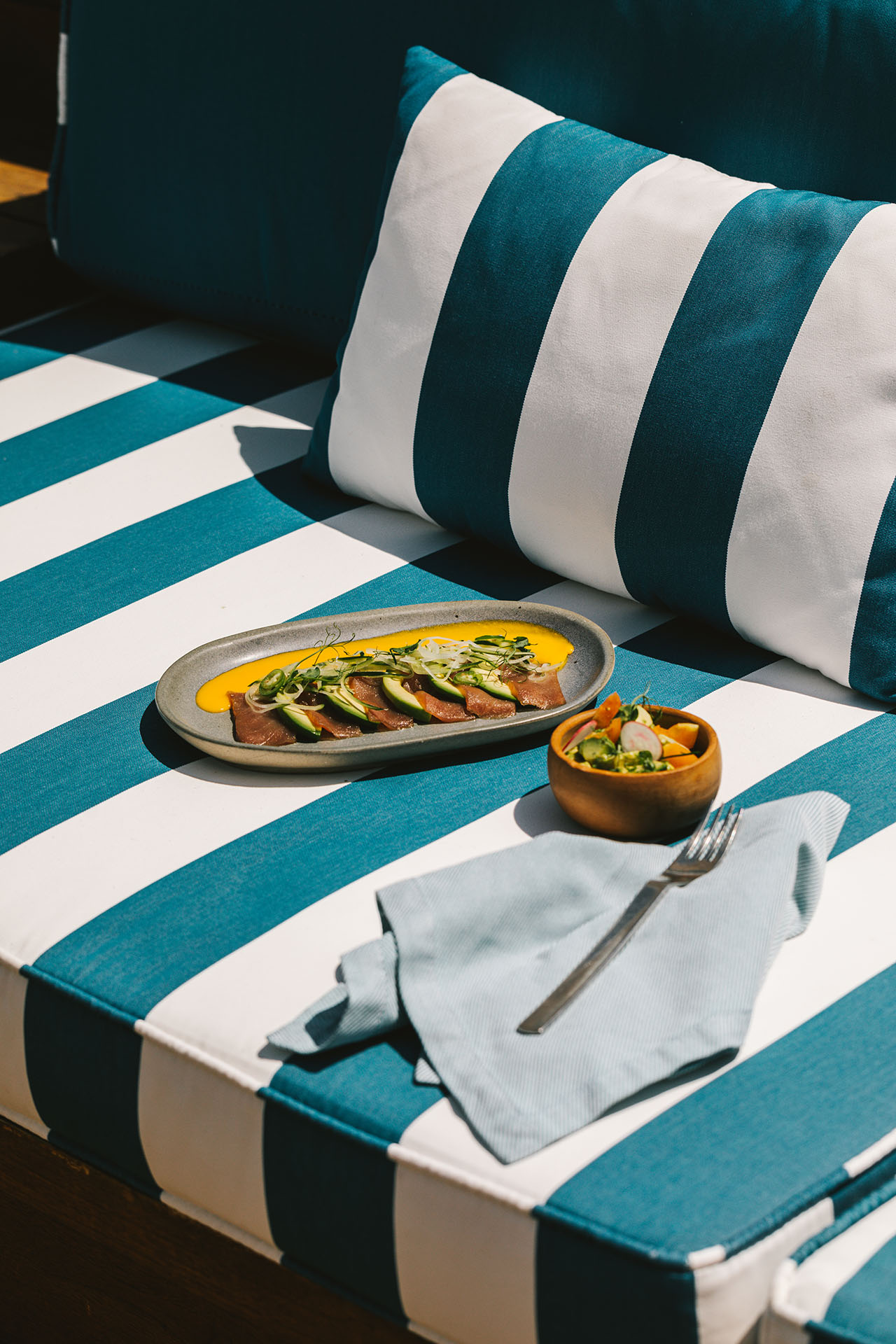 A plate of sliced meat with sauce and greens, and a small bowl of salad on a blue and white striped cushion with a napkin and fork.