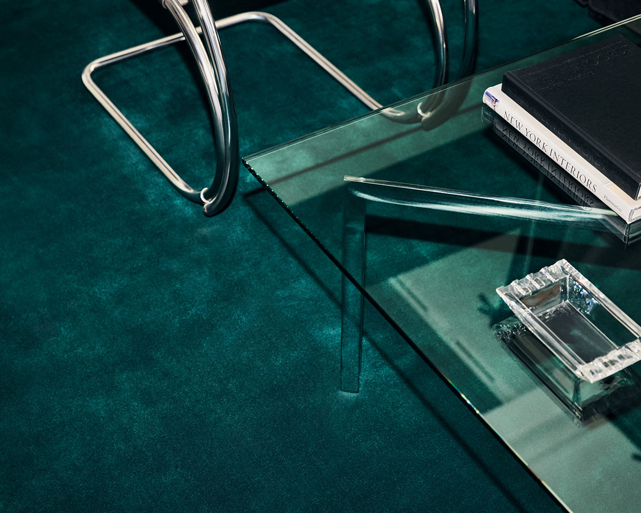 Dos libros y un cenicero de cristal rectangular se encuentran sobre una mesa de café de cristal sobre una alfombra verde azulado; Cerca se puede ver una pata de silla cromada.