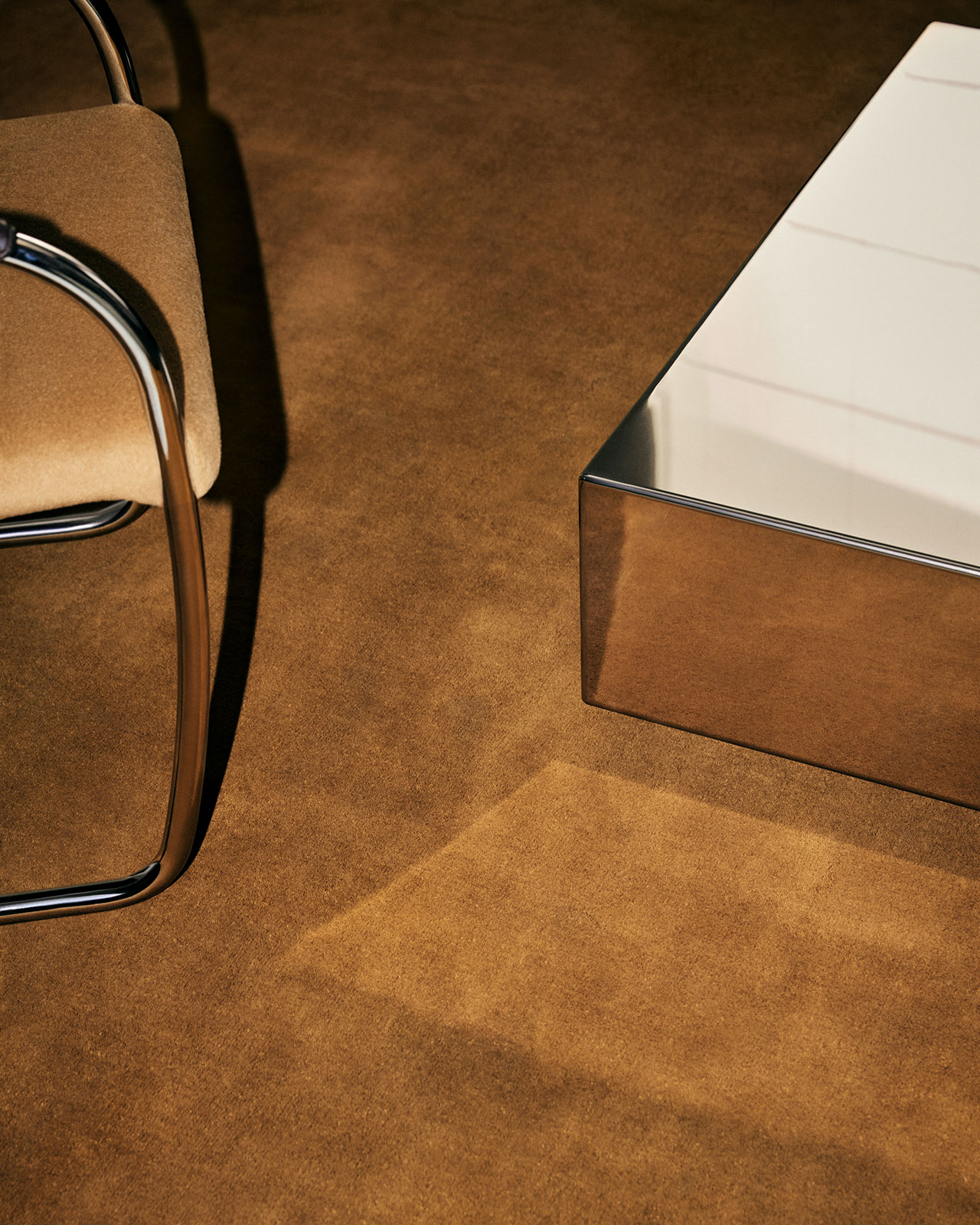 Una silla con estructura cromada y tapicería color canela se encuentra junto a una mesa de café rectangular con una superficie reflectante, ambas cubiertas con una alfombra marrón.