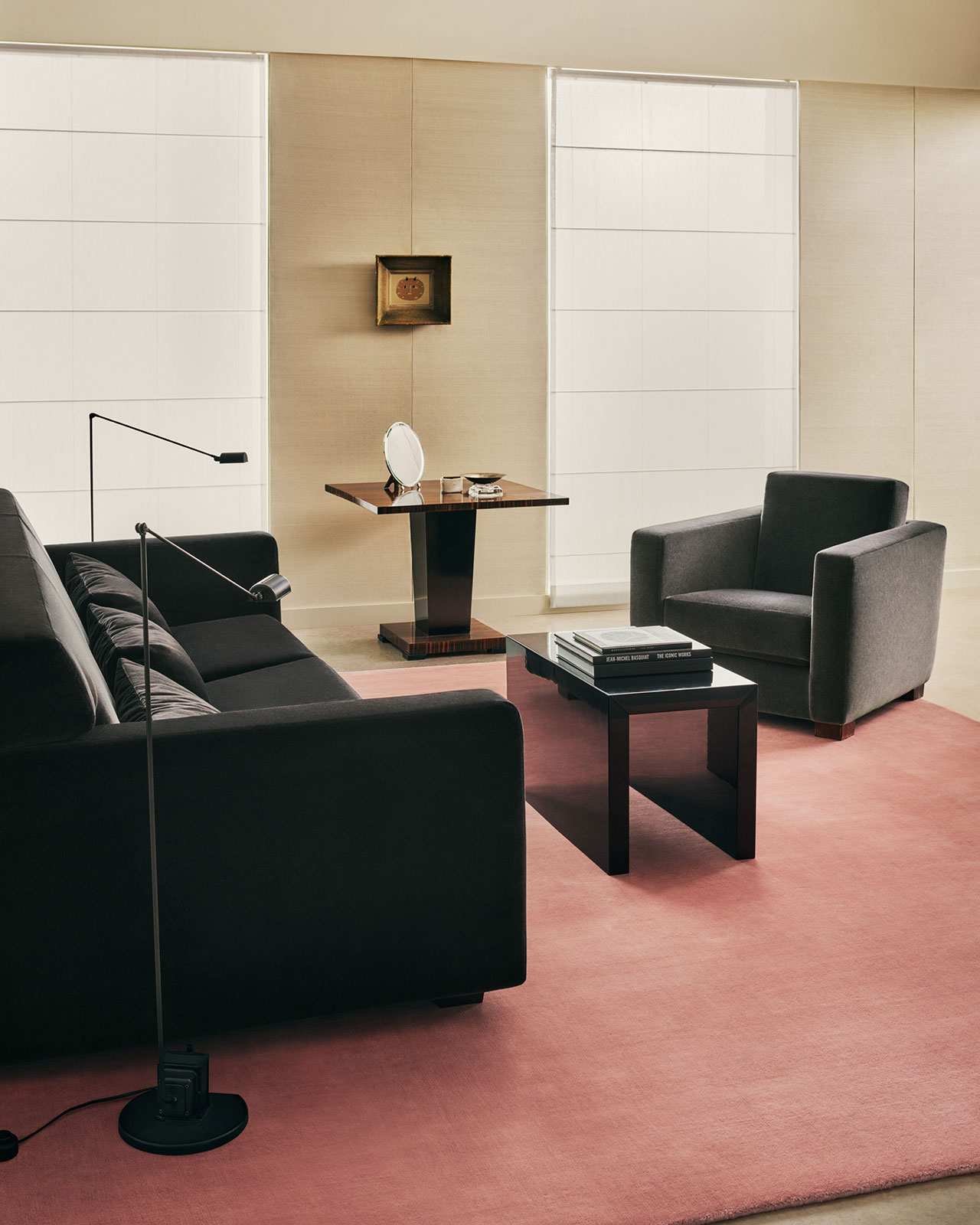 Salón moderno con sofá negro, sillón, mesa baja de cristal, lámpara de mesa, elementos decorativos en la mesa auxiliar y alfombra rosa sobre suelo beige.
