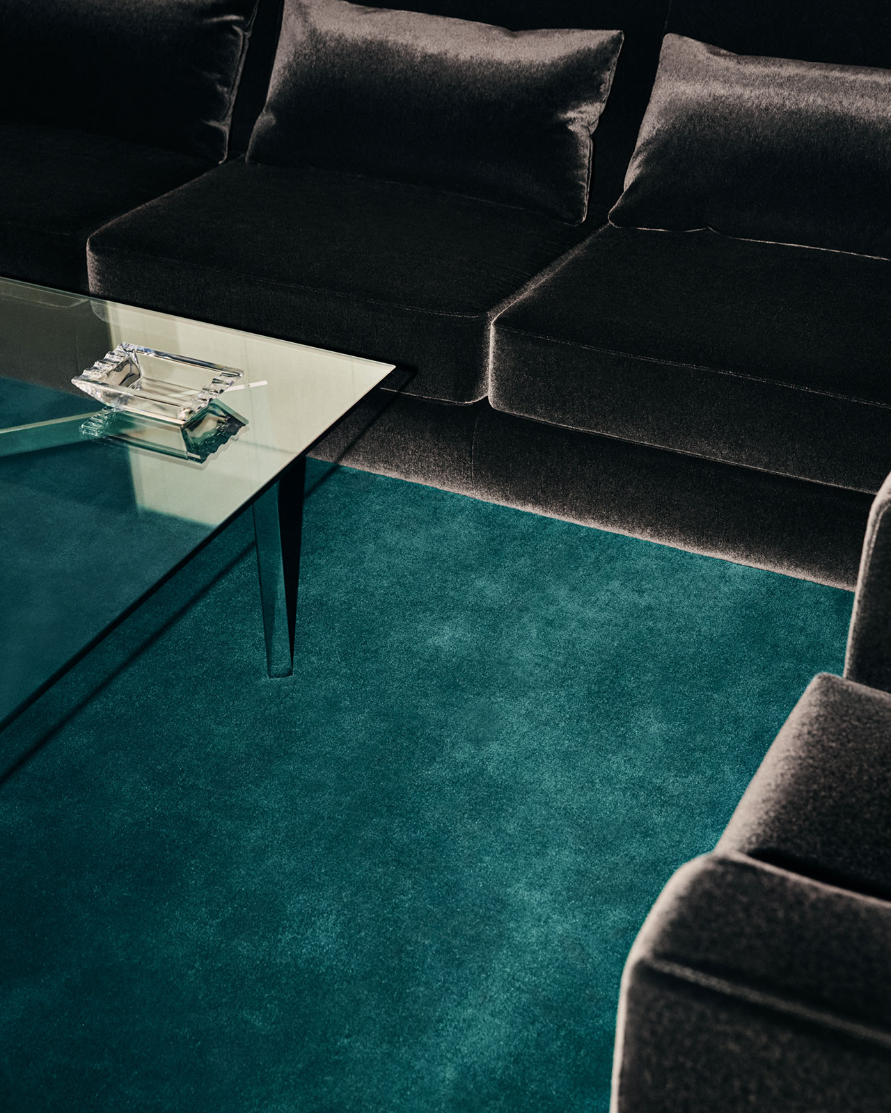 Salón moderno con mesa de centro de cristal, sofá de terciopelo oscuro y alfombra verde azulado. Hay un cenicero y un encendedor sobre la mesa.