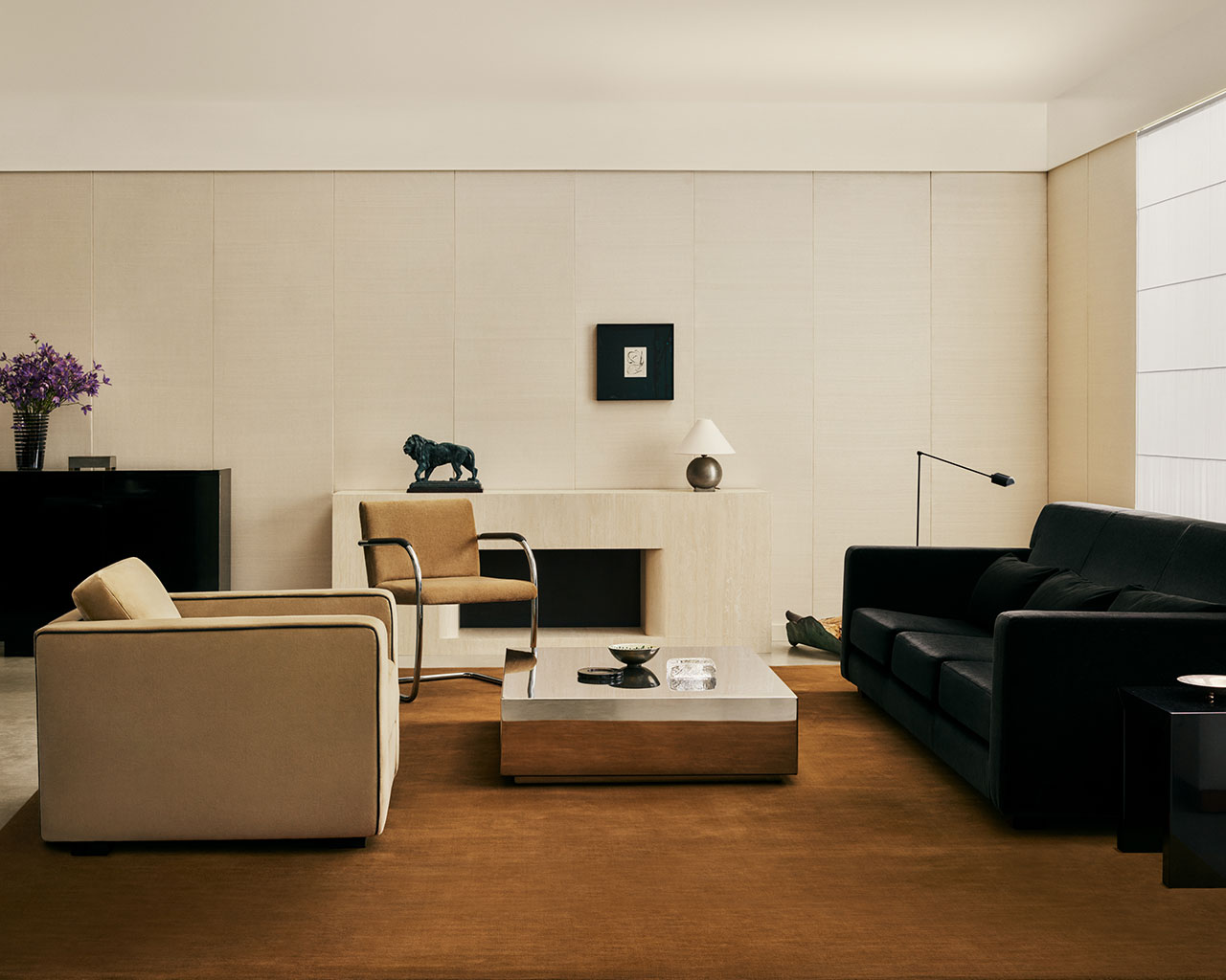 Salón moderno con sillón beige, sofá negro, mesa baja cuadrada, alfombra marrón, arte en la pared, lámpara de mesa, escultura y un jarrón de flores violetas en el aparador.