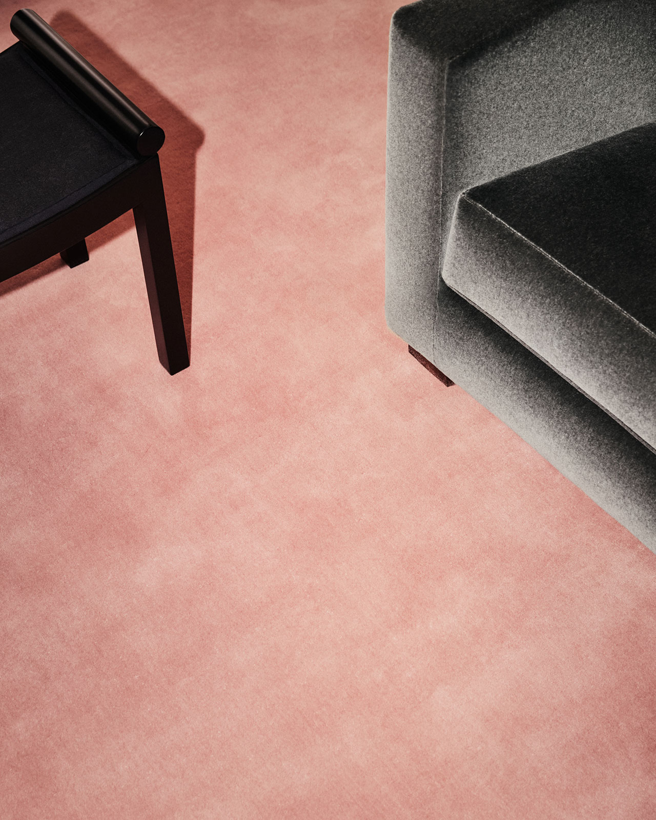 Bajo luces brillantes, un sillón tapizado en gris y una mesa auxiliar negra se encuentran sobre un piso alfombrado de color rosa claro.