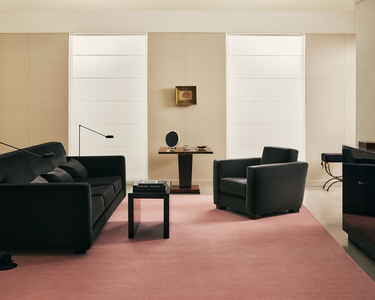 Salón moderno con sofá negro, sillón, mesa auxiliar y escritorio sobre alfombra rosa, con dos grandes ventanales y una pequeña obra de arte enmarcada en la pared trasera.