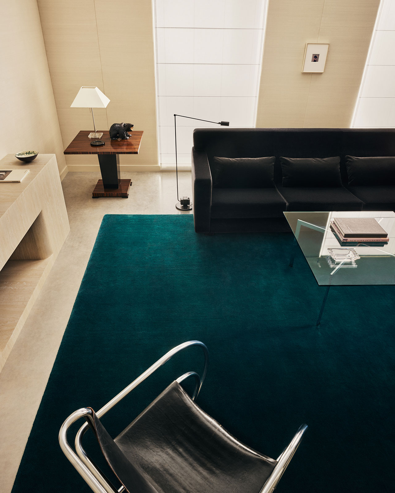 Salón moderno con sofá negro, mesa baja de cristal, alfombra verde azulado, elegante silla negra y mesa auxiliar con lámpara, frente a una ventana alta y una pared neutra.
