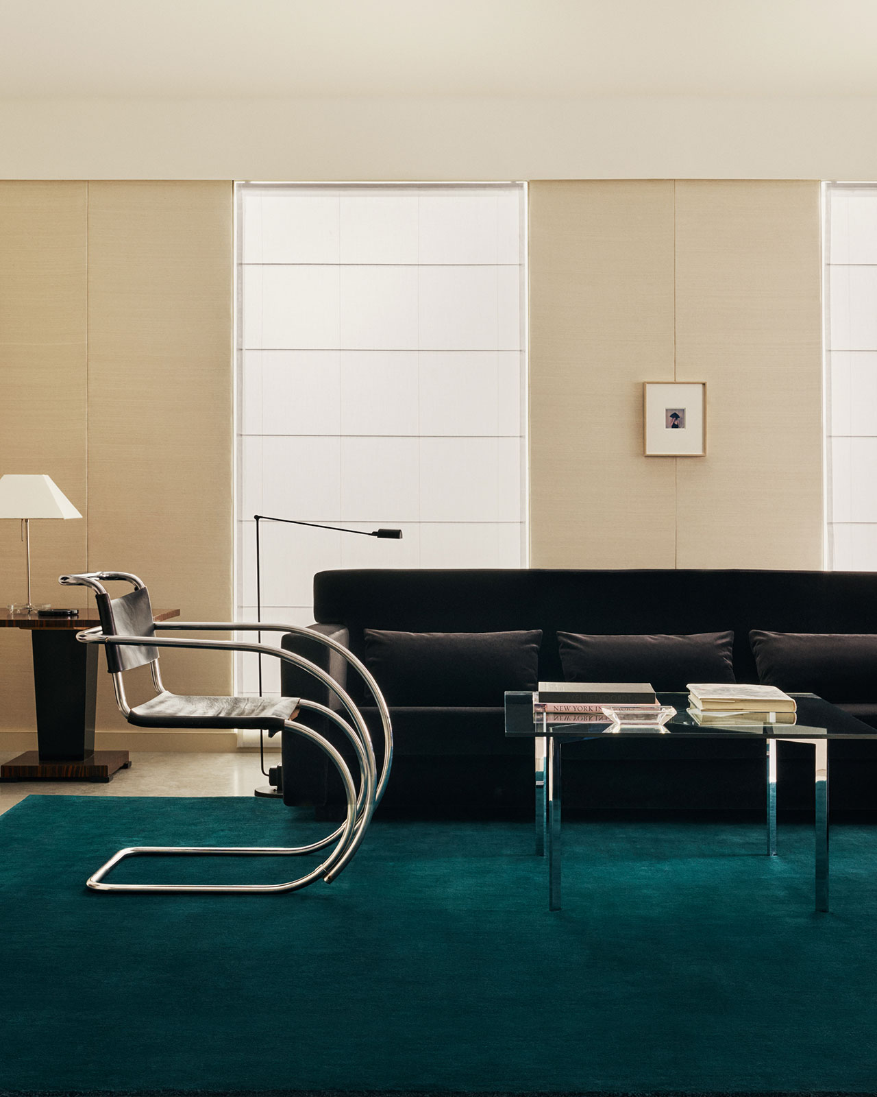 Salón moderno con sofá negro, mesa baja de cristal con libros, sillas cromadas, mesa auxiliar con lámpara y pared neutra con ventanas altas.
