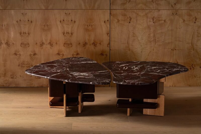 Two asymmetrical, dark marble coffee tables with irregular shapes and geometric wooden bases are placed on a light wooden floor against a wood-paneled wall.