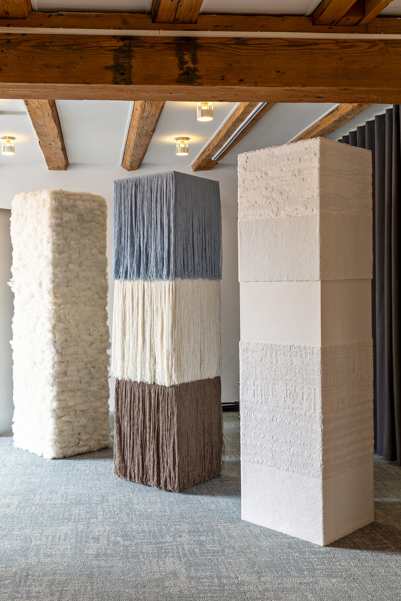 Three tall rectangular sculptures made of textured and fibrous materials stand in a room with exposed wooden ceiling beams and grey carpeting.