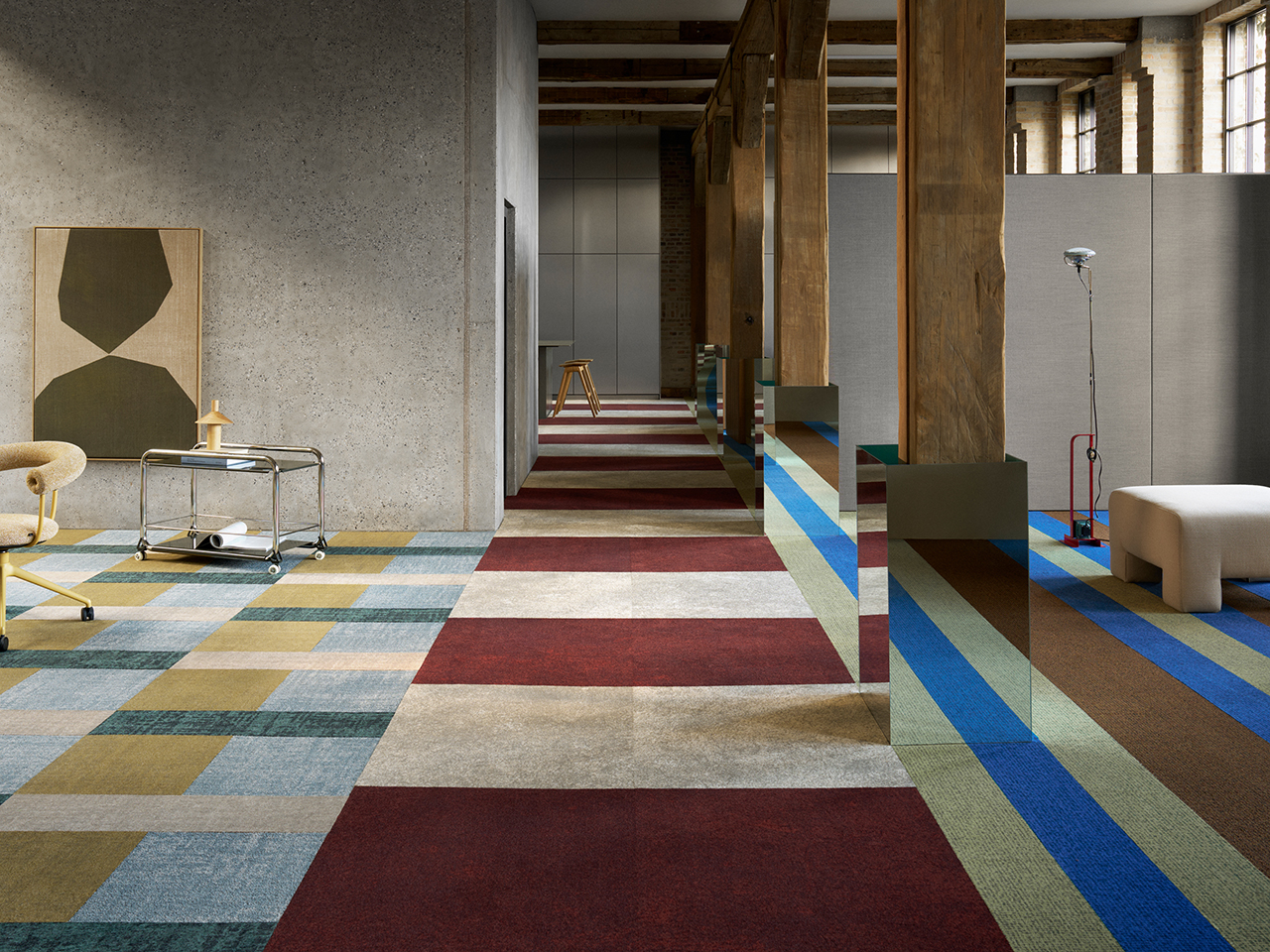 Modern interior space with exposed beams, concrete walls, and colorful geometric carpet tiles in red, blue, beige, and green. Minimalist furniture and abstract art are present.