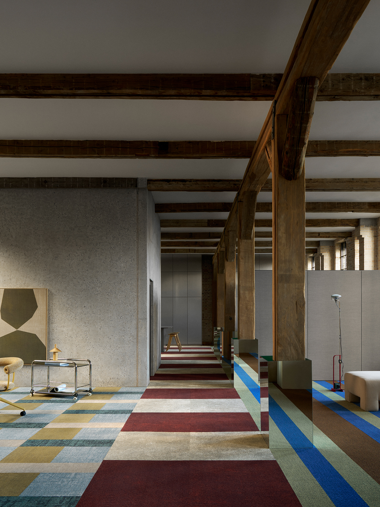 Modern office space with exposed wooden beams, a patchwork carpet in red, blue, yellow, and gray, concrete walls, and minimal furniture including a desk and lamp.