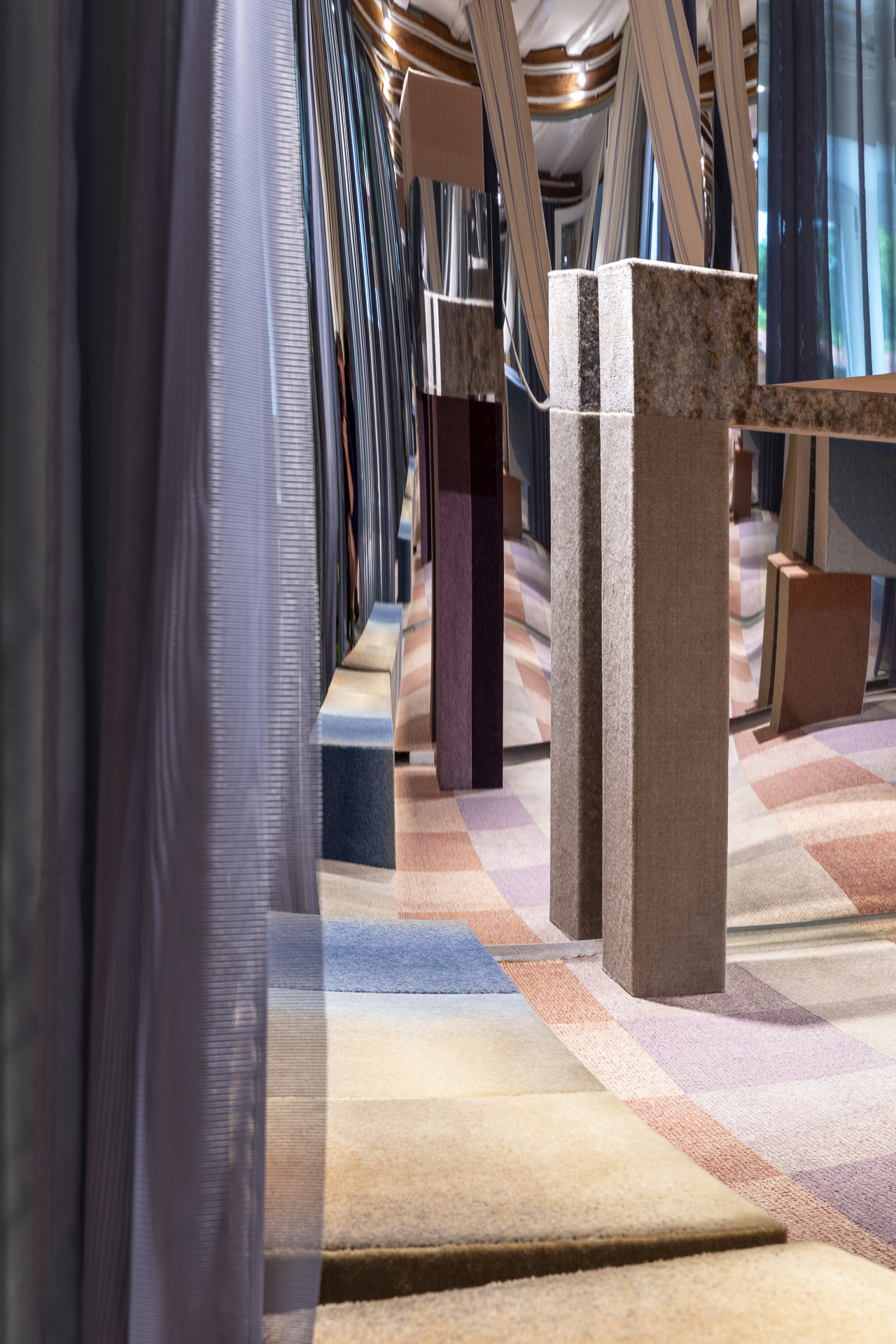 La habitación tiene altos pilares rectangulares cubiertos de tela y pisos de alfombras geométricas de colores, parcialmente separados por cortinas transparentes.