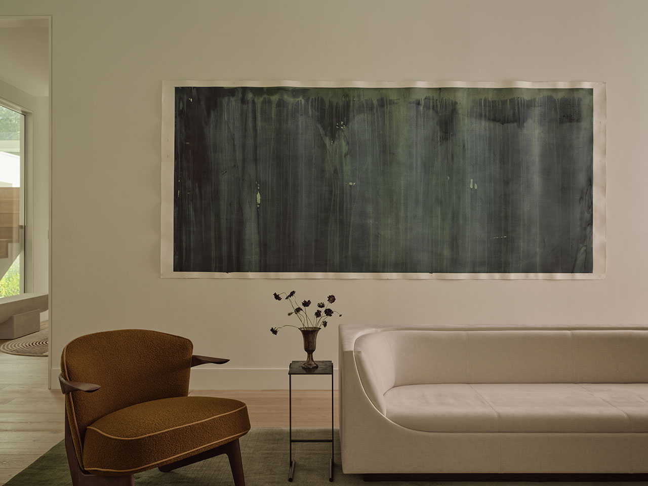 A modern living room with a brown armchair, white sofa, small side table with a vase of flowers, and a large abstract dark painting on the wall.