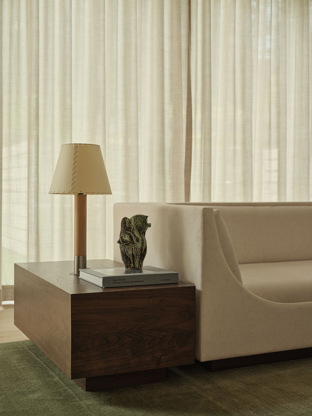 A wooden side table with a lamp, books, and a decorative sculpture sits next to a light-colored sofa in a room with sheer curtains and a green rug.